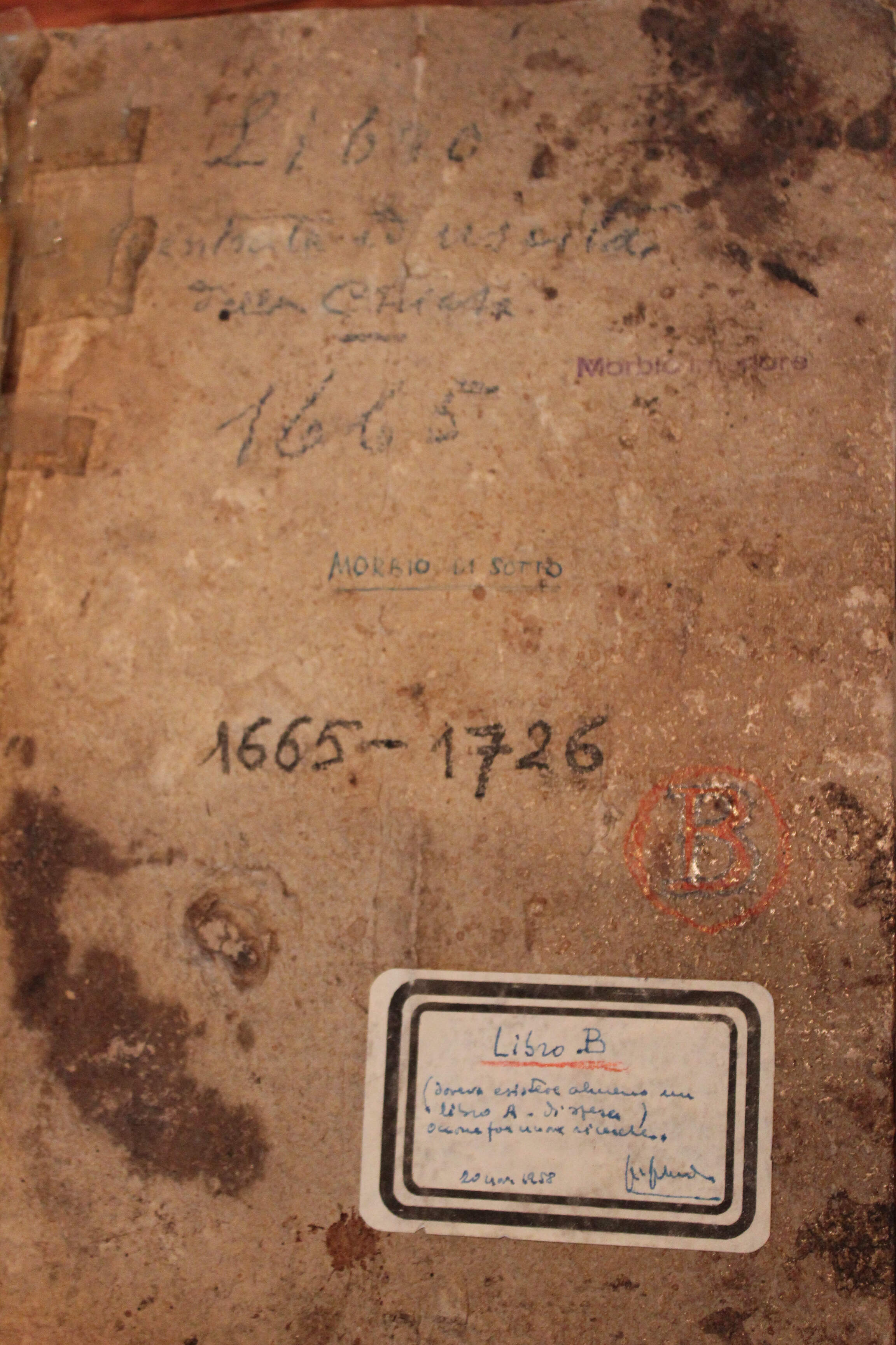A book cover made of stained and discolored parchment or leather, with handwritten inscriptions and markings in black and blue ink, a red stamp with the letter 'B', and a modern label with additional handwriting.